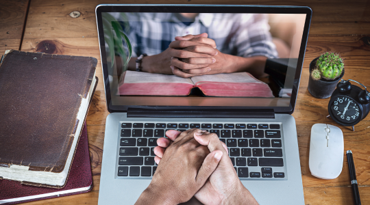 A person with folded hands in front of a laptop screen. An image of someone with bible is on screen signifying online bible resources